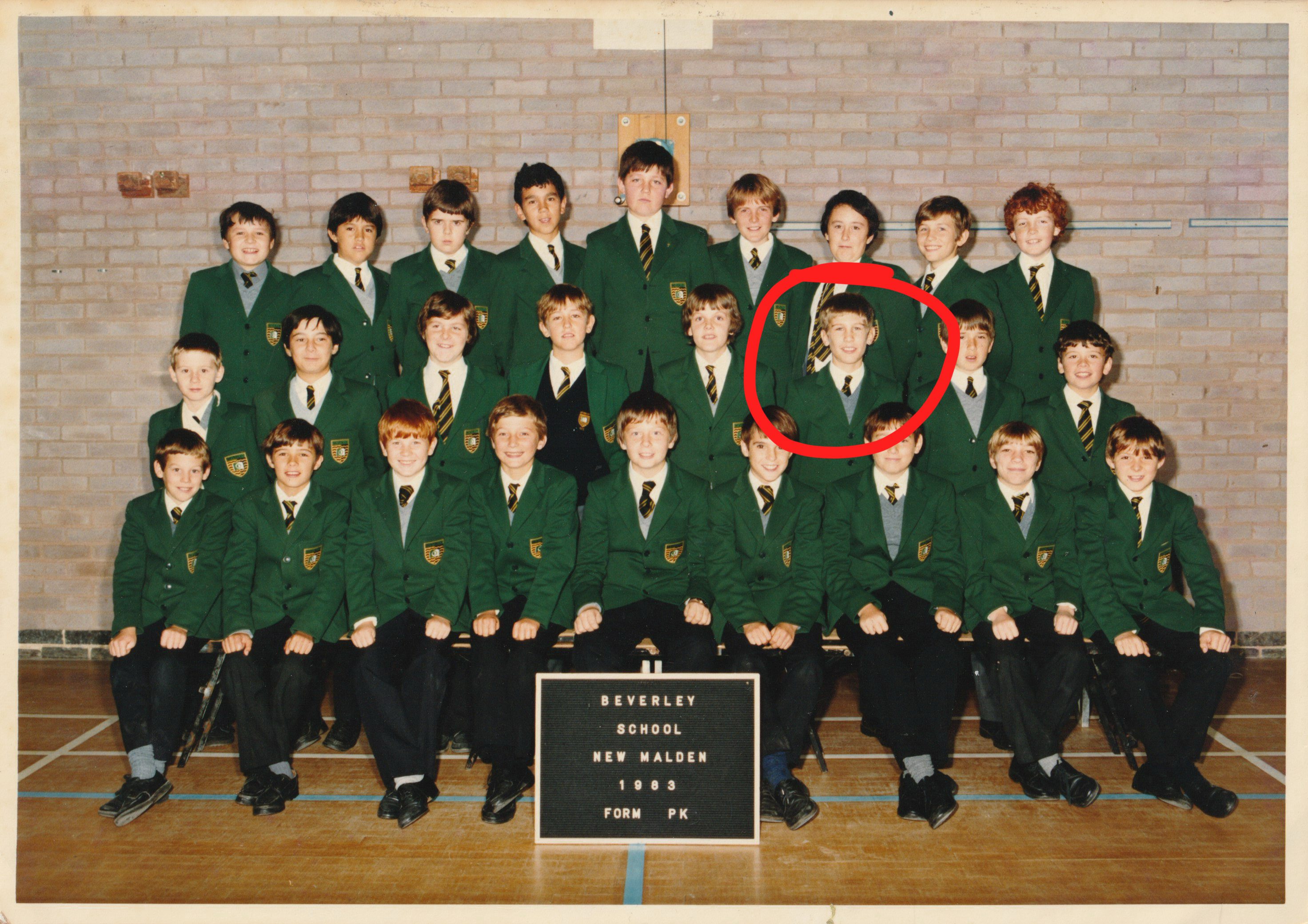 A colour school portrait of approximately 25 students, likely 10-12 years old, from Berverley School in New Malden, 1983, Form PK. The students are arranged in three rows in front of a brick wall, all wearing dark green school blazers, white shirts, and striped ties. A red circle is drawn around one boy in the second row.
