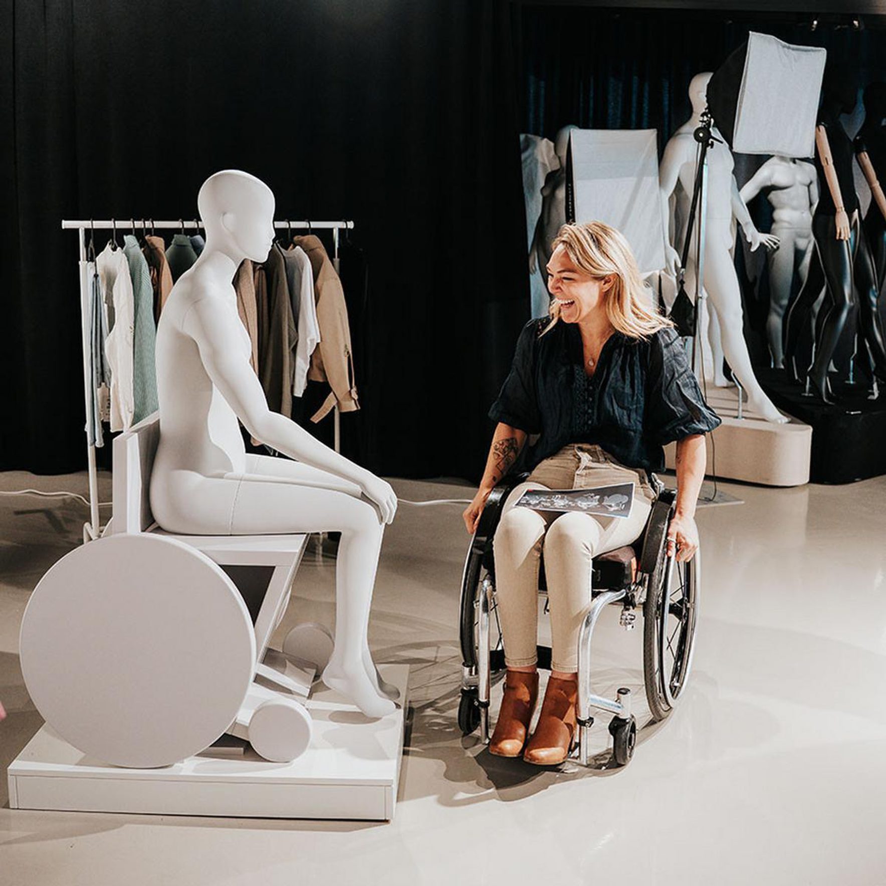 A smiling woman, identified as Sophie Morgan, sits in her manual wheelchair, facing a white Primark retail mannequin that is also seated in a stylized white wheelchair base. They are positioned in a clothing display area with racks of clothes and other mannequins in the background.