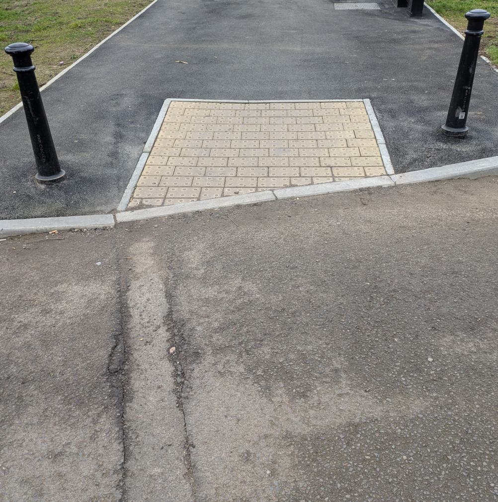 A dropped kerb (curb cut in the US) showing the transition between the road visible in bottom part of the image to the pavement (sidewalk in US) above it. The pavement is a tarmac and there is an area within it, closest to the road that looks like it is textured tiles for increased tactlessness. Black posts sit either side of the textured tiles.