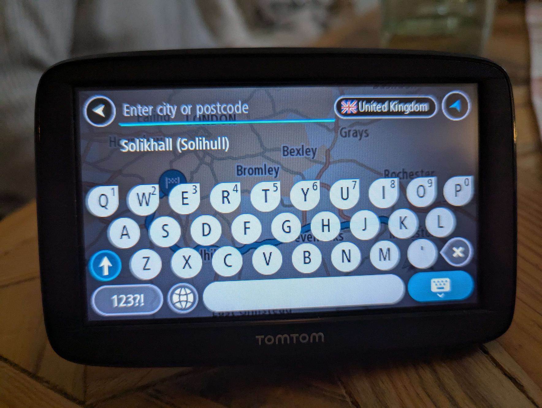 A touch screen TomTom device with various input elements including a key pad and a search field with placeholder text: Enter city or postcode. The placeholder text is pale blue overlaid on a map making it difficult to read.