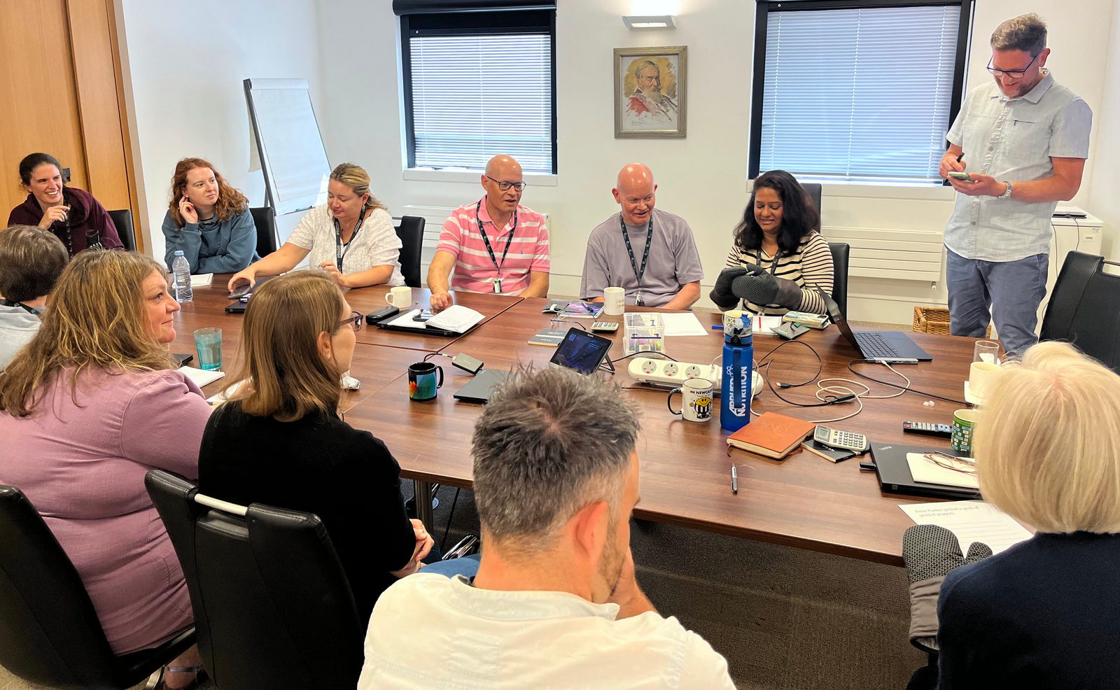 A photo of a diverse group of ten people gathered around a long wooden conference table in a bright meeting room. Laptops, mugs, and water bottles are scattered across the table. One man in a light blue shirt stands on the far right, and another man in a pink striped shirt is seated in the middle foreground, looking down at the table.
