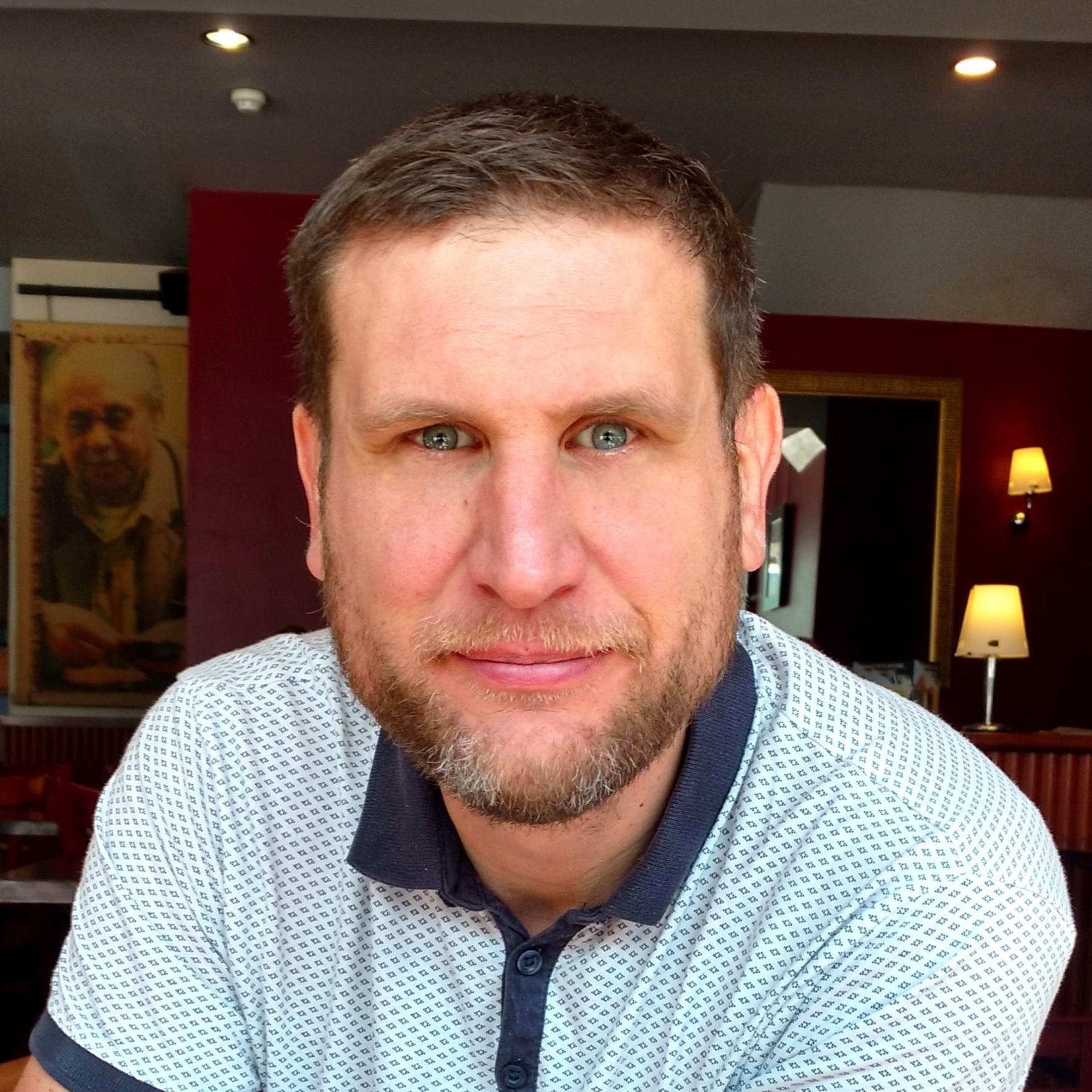 Simon Leadbetter is a white middle aged man with shortish medium brown hair and blue eyes. He has a short beard with (only) a few grey hairs.
